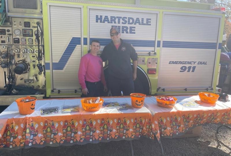 Two people pose by a fire truck, with a table featuring Halloween-themed decorations and candy buckets.