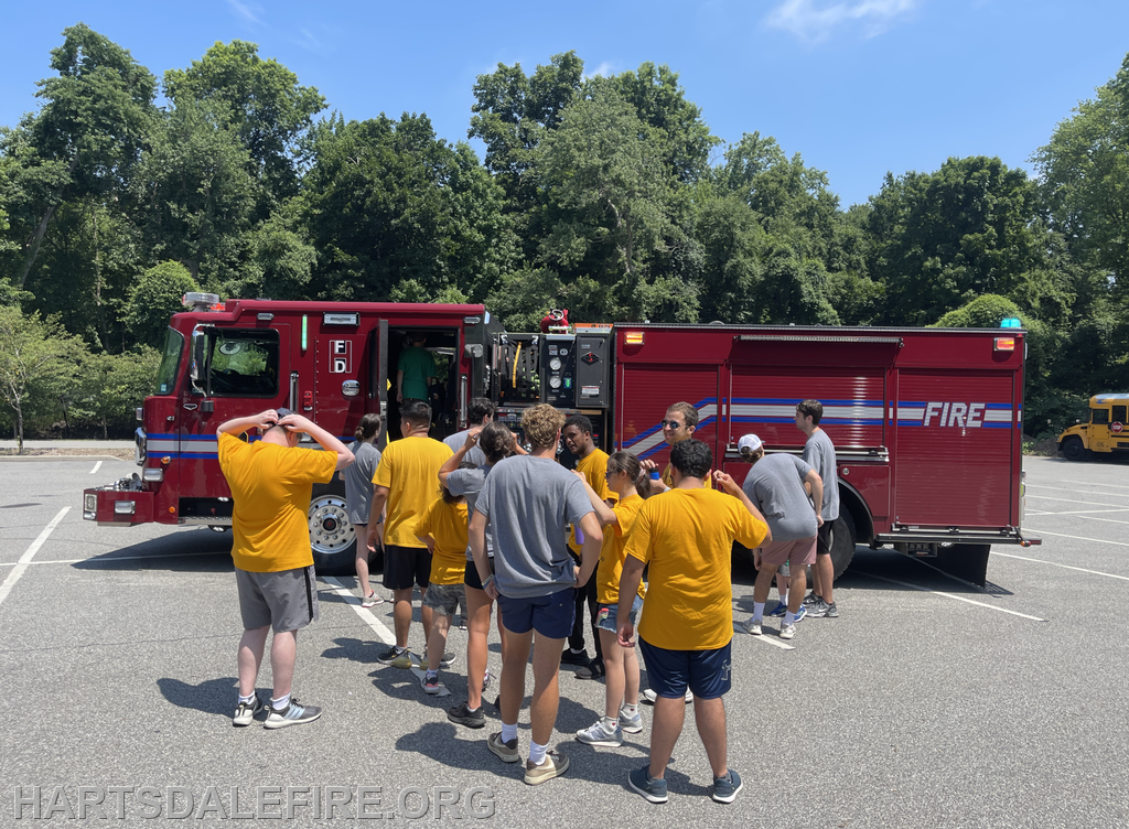 A group of people in yellow shirts gathers around a fire truck in a parking lot on a sunny day.