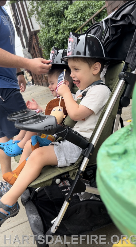 A young child in a stroller wears a firefighter hat, smiling and holding a toy, while another child sits nearby.