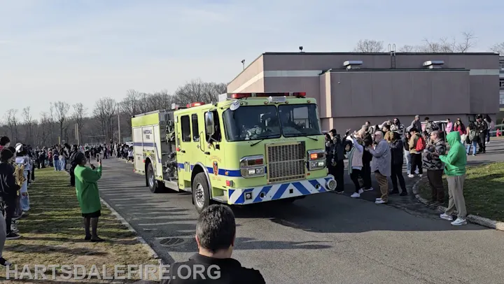 A bright green fire truck drives by a crowd of people, some taking photos, at an outdoor event near a building.