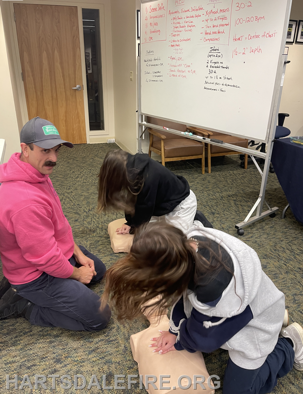 Two individuals are practicing CPR on mannequins while a trainer provides guidance in a classroom setting.