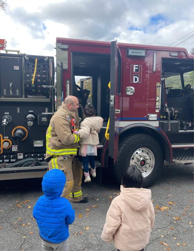 A firefighter helps a child into a fire truck while two other kids watch, showcasing a community event.