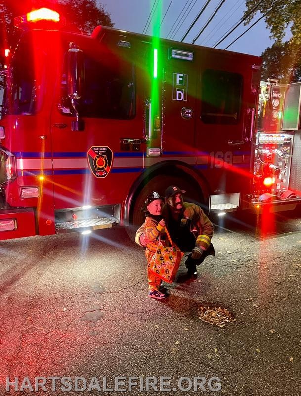 A firefighter poses for a photo with a child dressed as a fire chief in front of a fire truck, illuminated by red lights.
