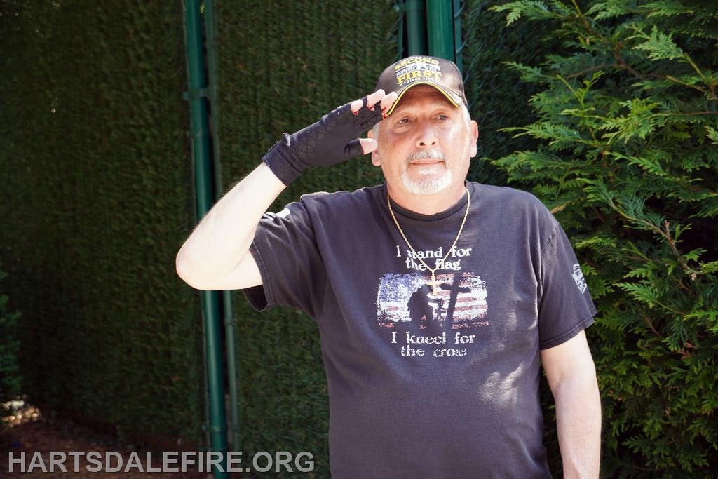 A man saluting, wearing a cap and a t-shirt with a flag design and message.