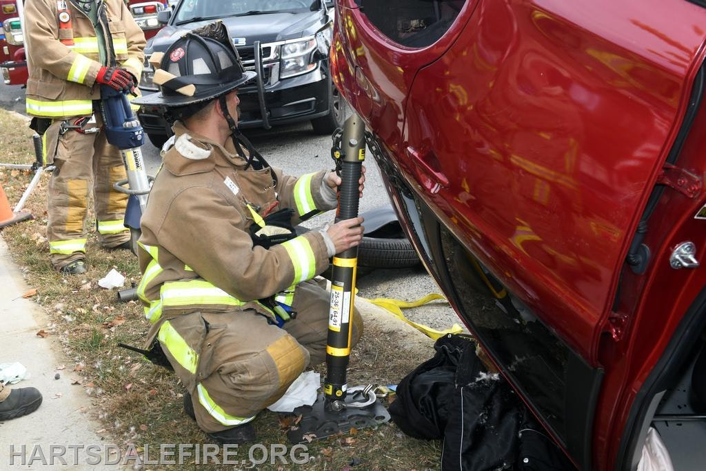 Firefighters are using rescue equipment to assist at the scene of a vehicle incident, focusing on a red car.