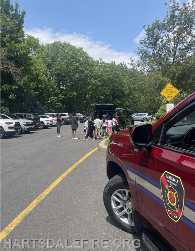 A group of children gather near a parked SUV while a fire vehicle is in the foreground under a clear blue sky.