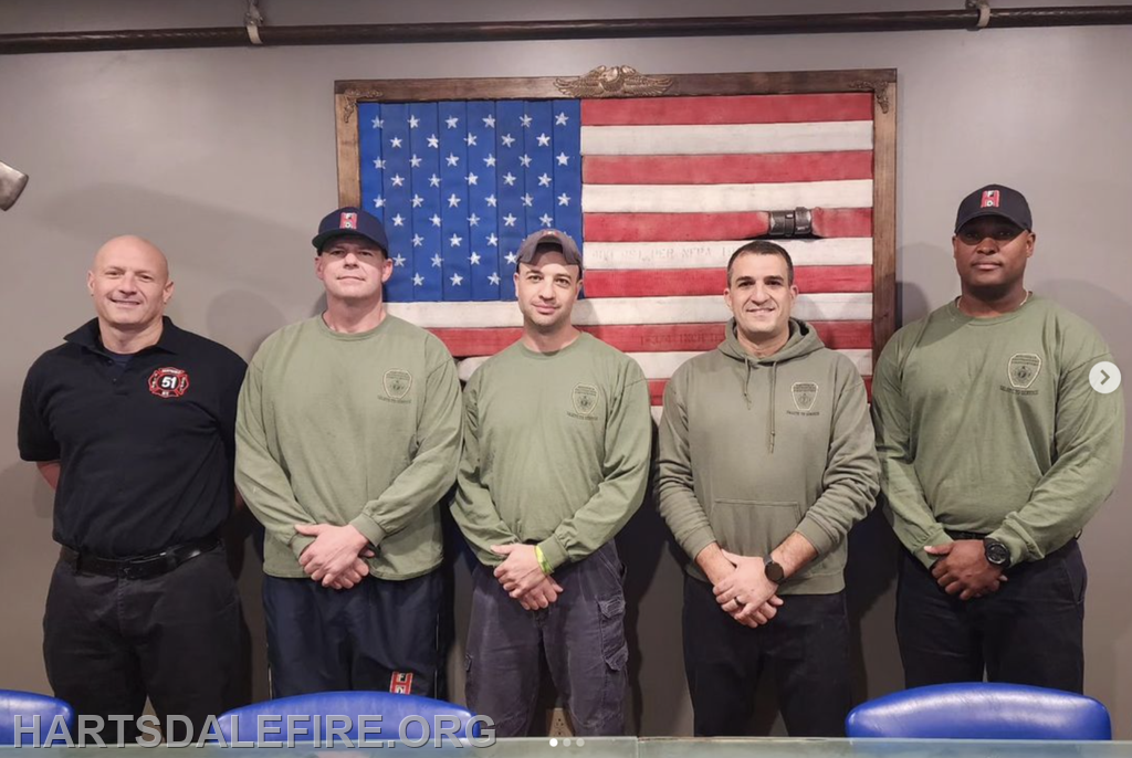 A group of five individuals, likely firefighters, stand together in front of an American flag backdrop.