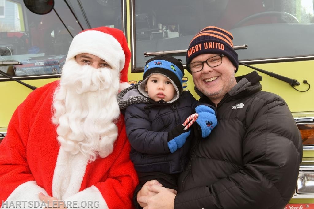 A person dressed as Santa sits beside a man holding a child with a candy cane, in front of a yellow vehicle.