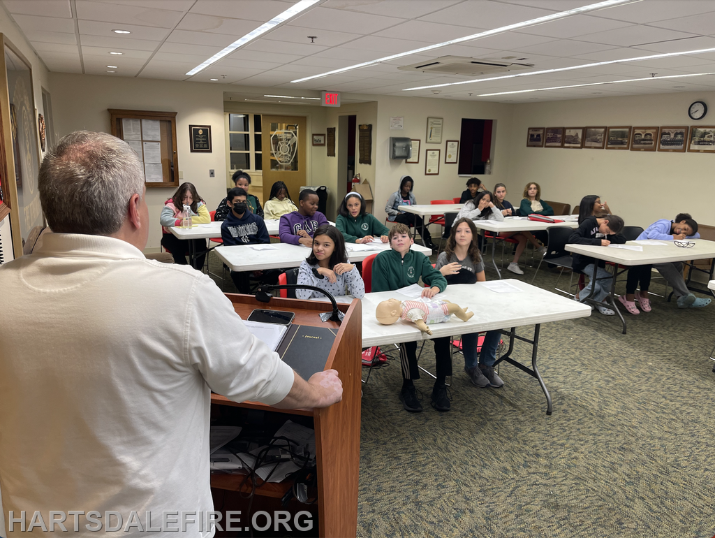 A person is lecturing a group of students in a classroom with a CPR dummy on a table.