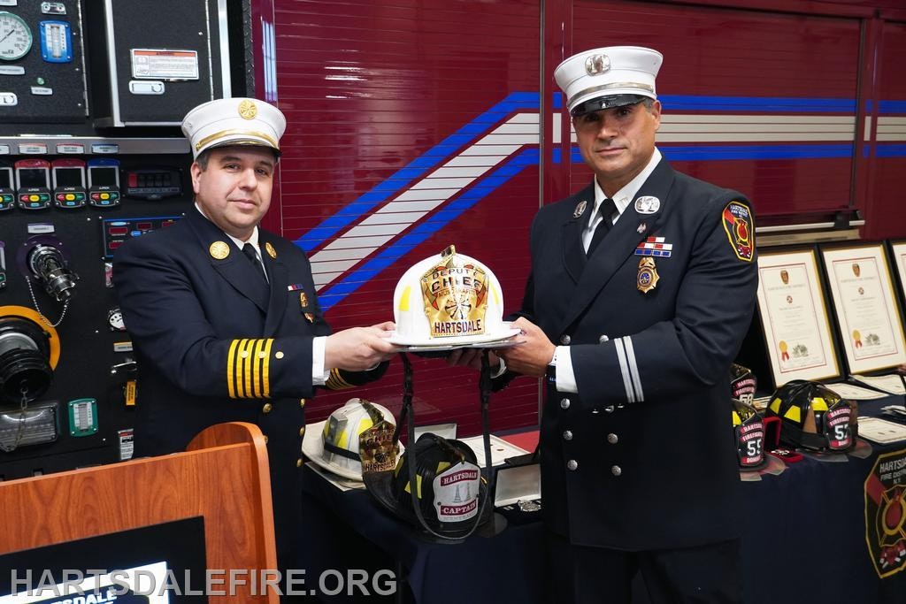 Two firefighters in uniform holding a white fire chief helmet in front of a firetruck and framed certificates.
