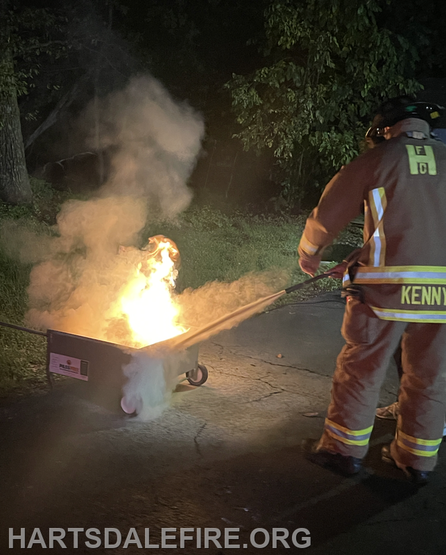 A firefighter, dressed in gear, uses a tool to manage a fire in a wheelbarrow at night, producing smoke and flames.