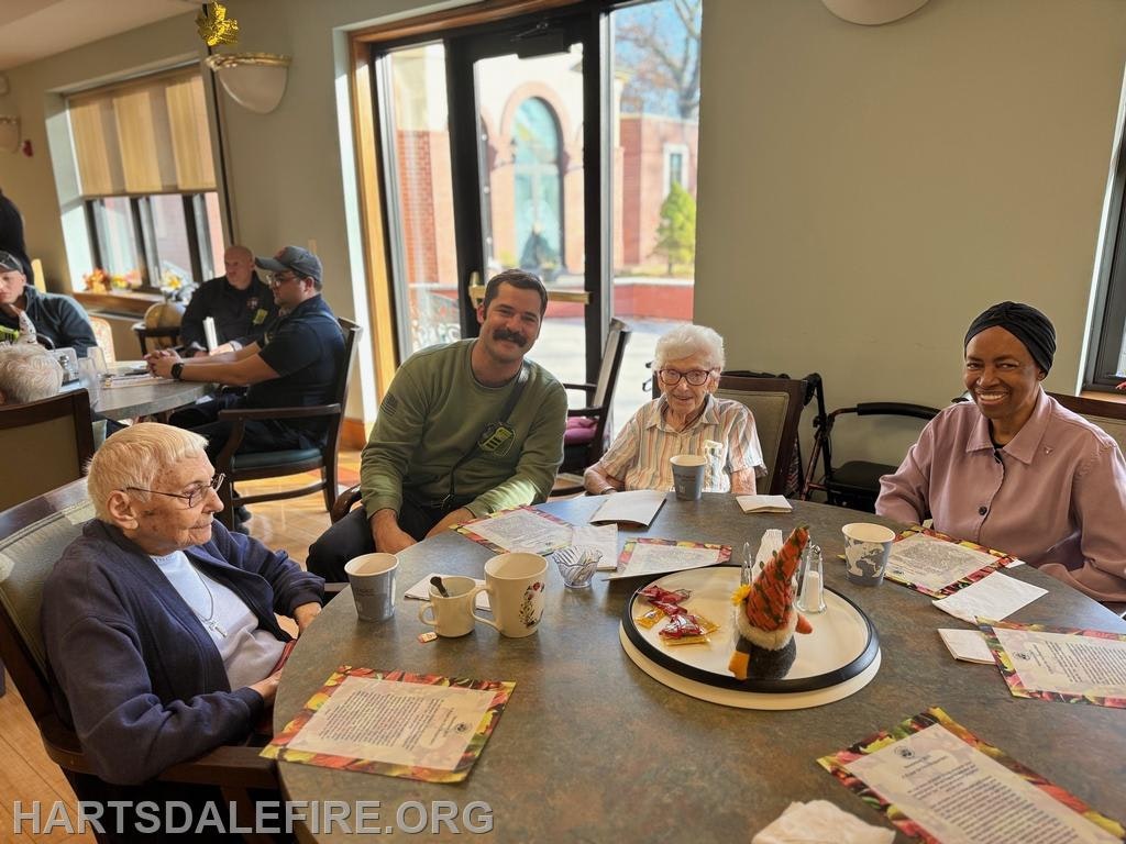 A group of four people, including seniors and a man, are gathered around a table in a cozy dining area, enjoying drinks and chatting.