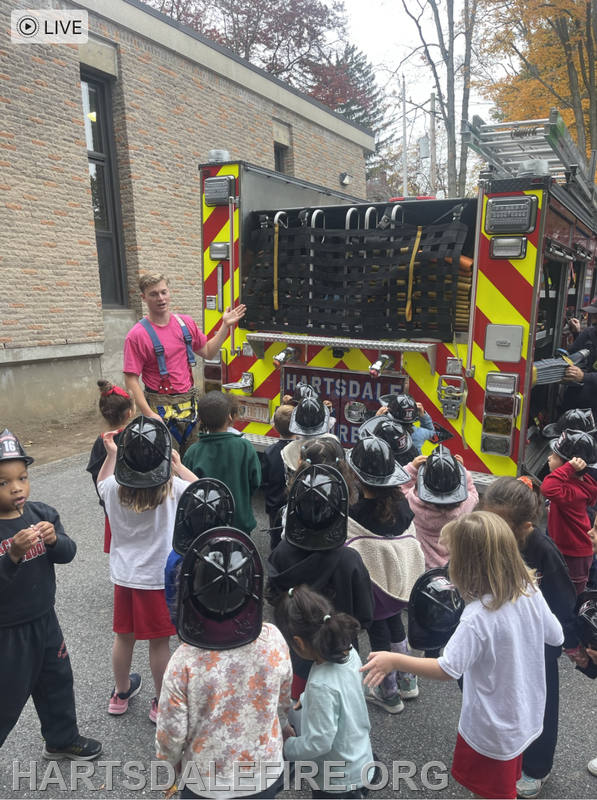 A firefighter in pink is showing kids in helmets about a fire truck during a safety demonstration.