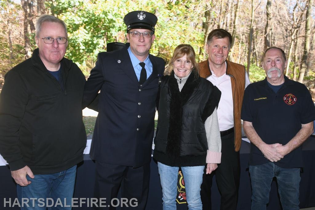 A group of four people, including a firefighter in uniform, poses outdoors in a wooded area, likely at a community event.