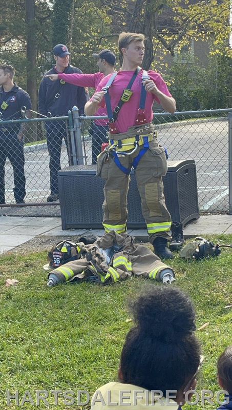 A firefighter in gear demonstrates equipment while others observe nearby, showcasing fire safety training or event.