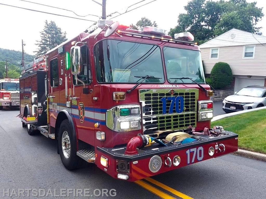 A red fire truck marked 170 is parked on the street with hoses attached, in a suburban neighborhood.