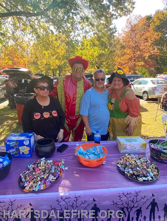 A group of people in costumes at a festive table with candy and treats, surrounded by autumn trees and parked cars.