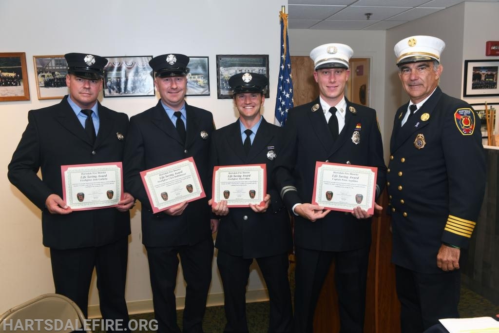Five uniformed firefighters holding certificates in an indoor setting, with an American flag in the background.