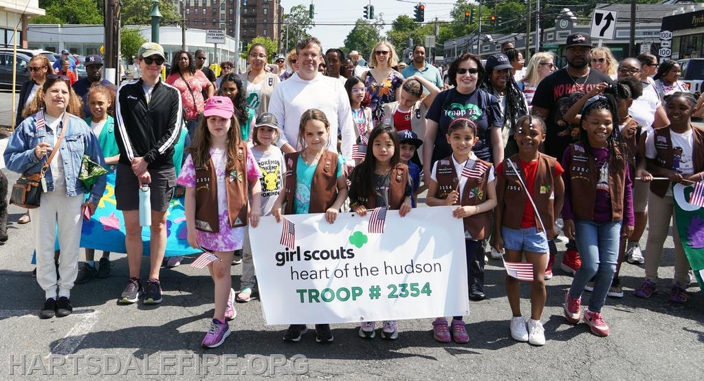 A Girl Scout troop in uniform holds a banner, "Girl Scouts Heart of the Hudson Troop #2354," in a parade setting.