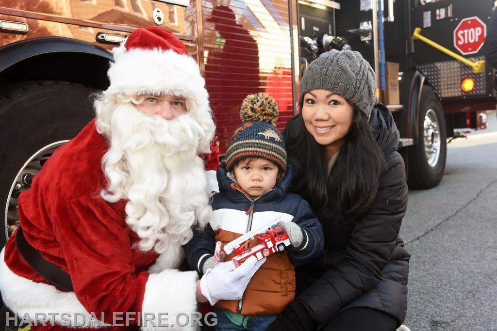 Santa poses with a woman and a young boy holding a toy fire truck, set against a backdrop of a fire truck.