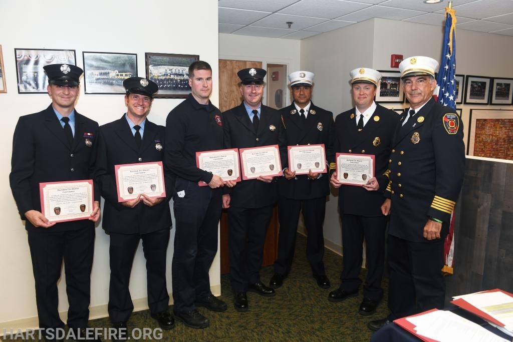 Group of firefighters in uniform holding certificates, standing indoors with framed photos and an American flag in the background.