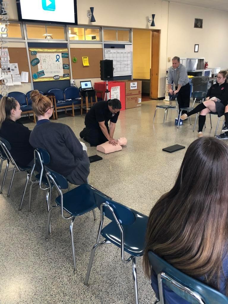 A person demonstrates CPR on a dummy to a seated group in a classroom setting.