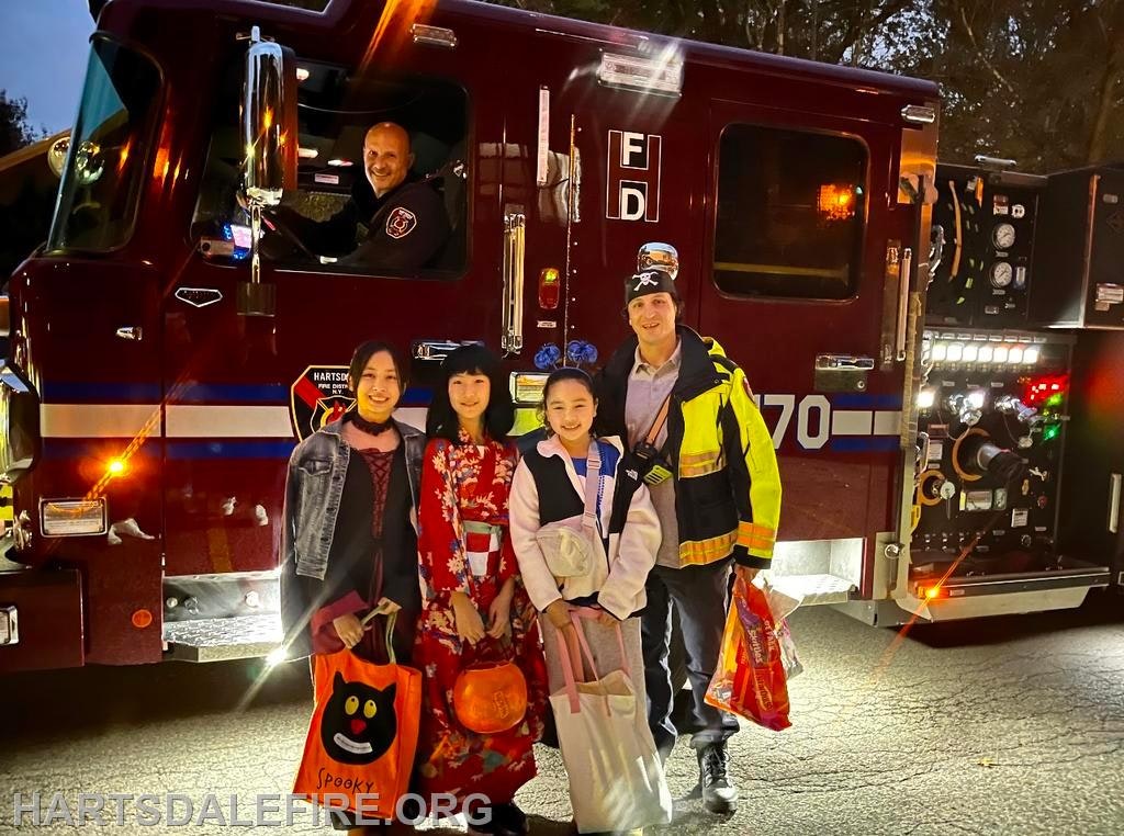 A fire truck with a firefighter, alongside four children in costumes holding bags, likely during a Halloween event.