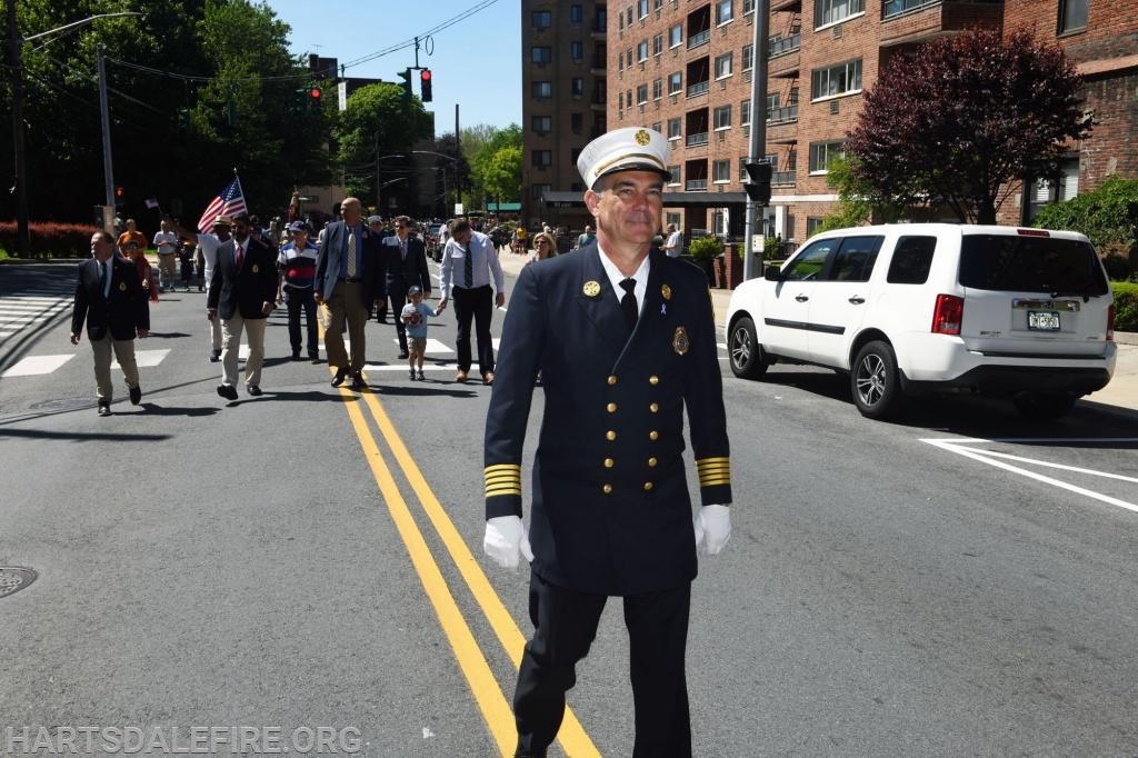 A uniformed fire chief leads a group in a parade on a sunny street, with people holding American flags.