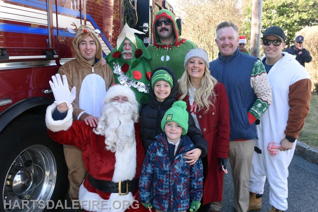 A festive group poses with Santa, wearing holiday costumes, near a fire truck, capturing a cheerful holiday spirit.