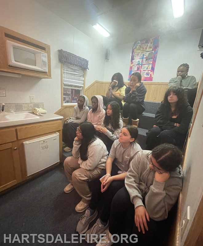 A group of young women is sitting on steps, attentively watching something on a screen in a cozy indoor setting.