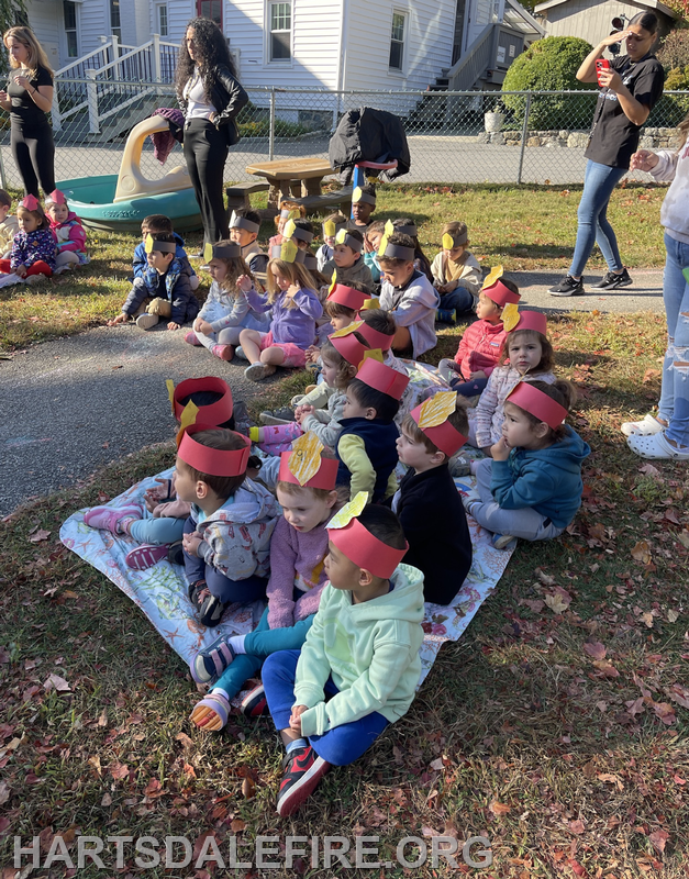 A group of children wearing colorful hats sits on a blanket enjoying a sunny outdoor activity with adults nearby.