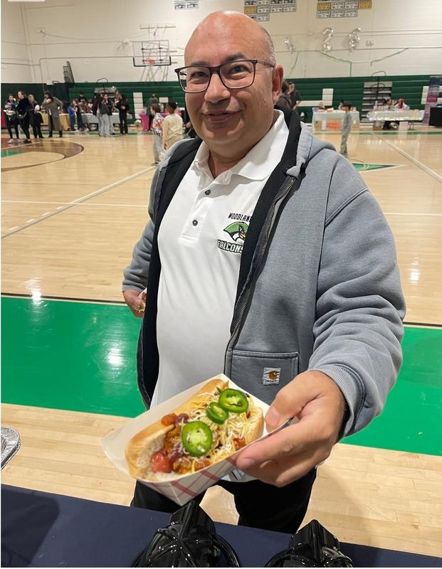 A smiling man in a gym holding a hot dog topped with jalapeños, surrounded by people and event tables in the background.