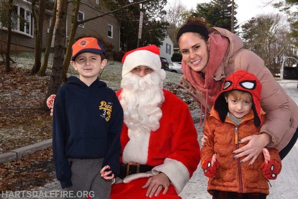 A person dressed as Santa Claus with a family, including two children holding candy canes, posing outdoors on a winter day.