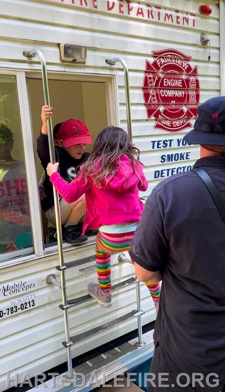 A child climbs a ladder into a fire safety trailer with the Fairview Fire Department logo.