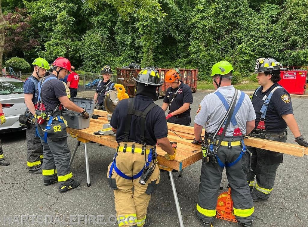 Firefighters in gear using tools and wood on a table outdoors.
