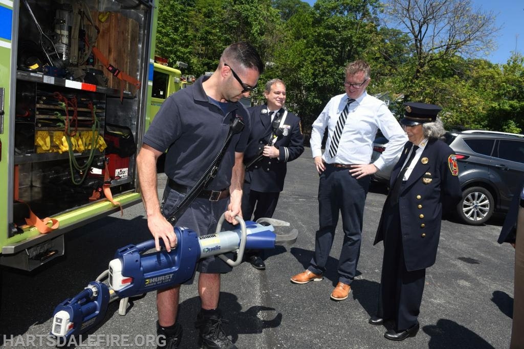 A firefighter demonstrates a rescue tool to onlookers by an open fire truck.