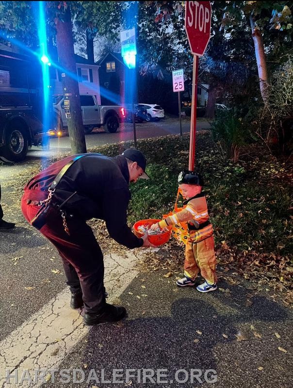 A firefighter is happily giving candy to a young child dressed as a firefighter during a nighttime event.