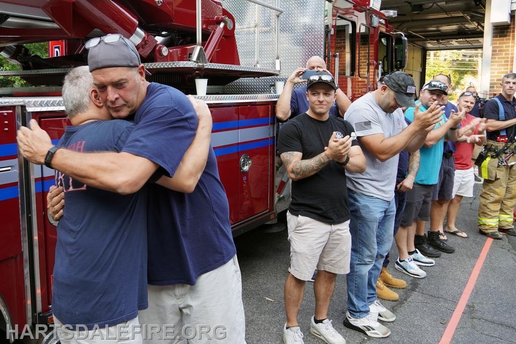 Two men hug near a fire truck while others stand nearby, clapping and observing the warm moment.