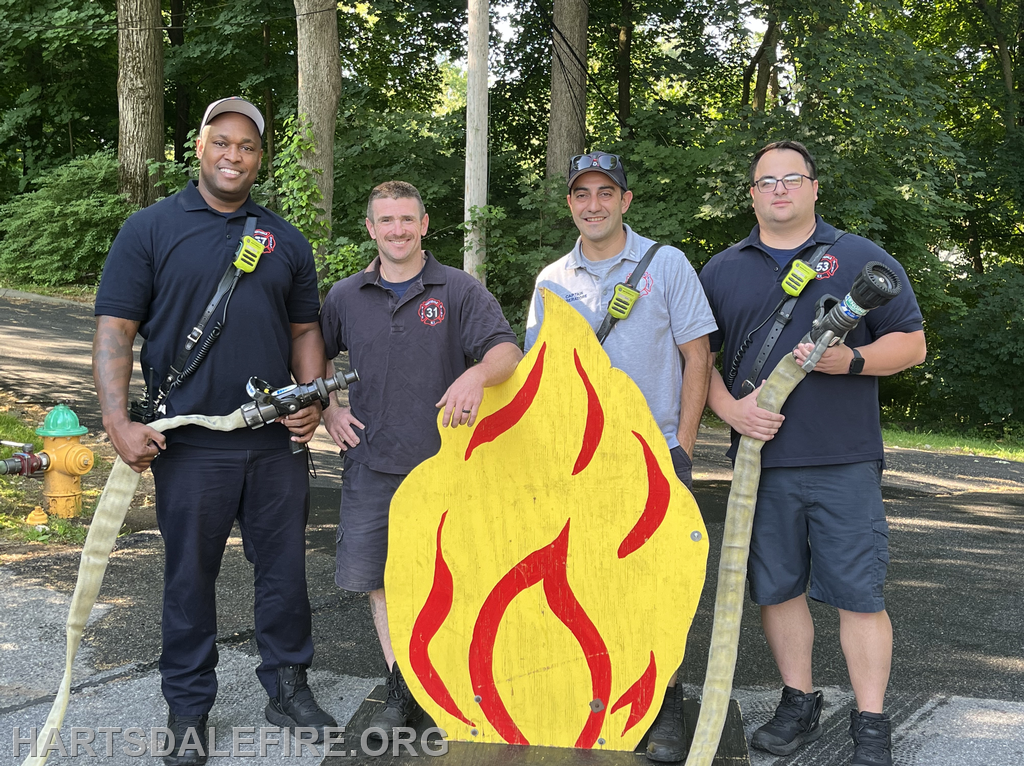 Four firefighters with hoses stand around a wooden flame cutout, smiling outside near trees and a fire hydrant.
