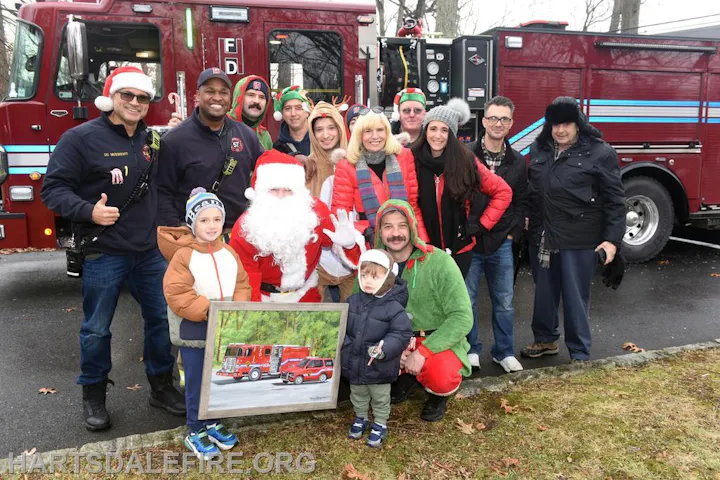 A festive group gathers around Santa and a fire truck, celebrating the holidays with joyful spirit and a painting.