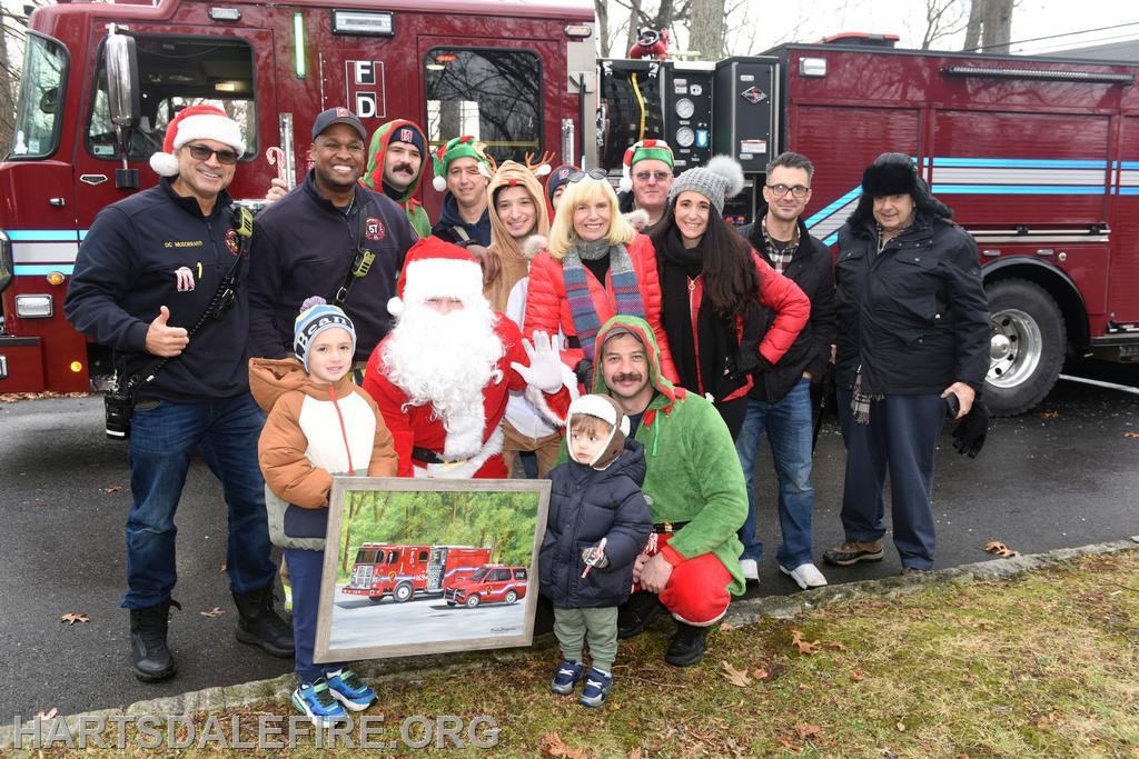 A festive group gathers around Santa and a fire truck, celebrating the holidays with joyful spirit and a painting.