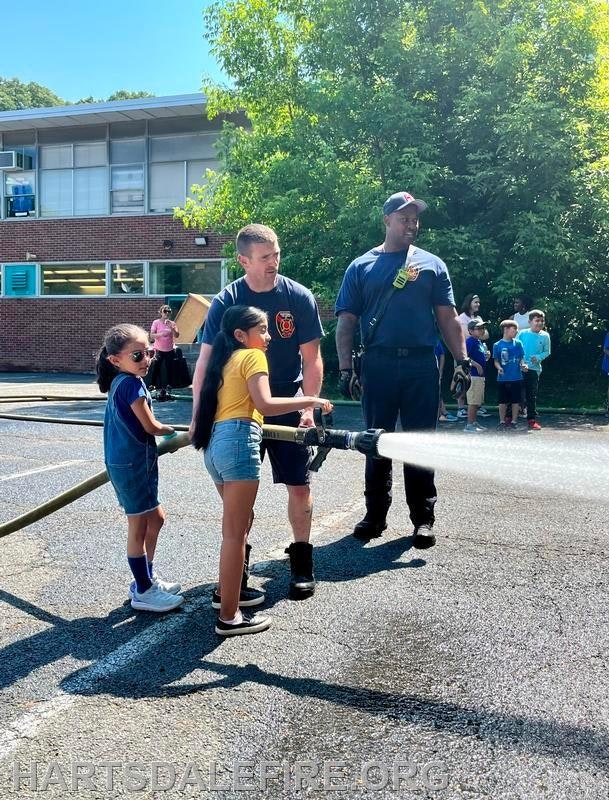 Children, with help from firefighters, spray water from a hose outside a building, while others watch.