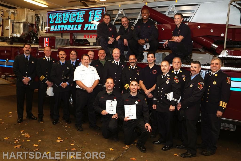 A group of firefighters poses in front of a fire truck, celebrating an achievement or award in a garage setting.