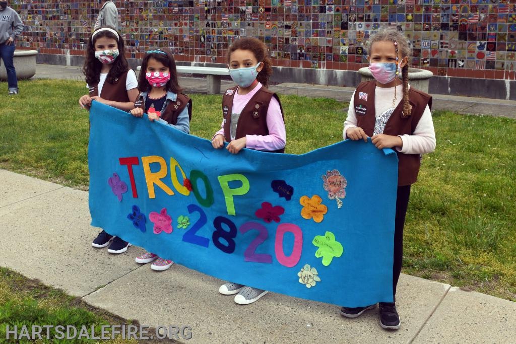 Four children in brown uniforms hold a colorful banner reading "TROOP 2820," standing outdoors on grass with masks on.