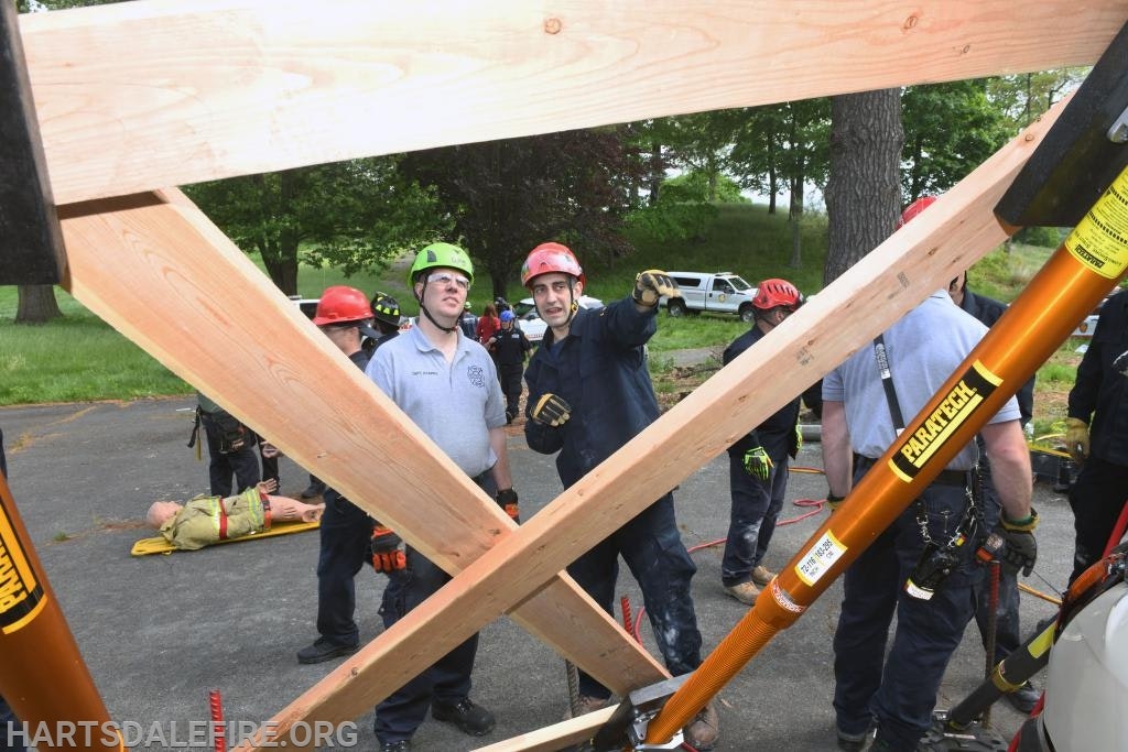 Rescue team practicing with wooden beams and equipment; mannequin on ground, group wearing helmets.