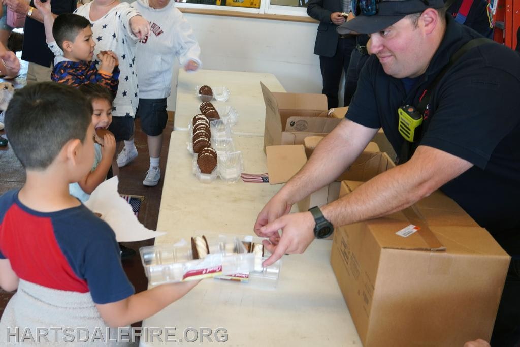 People at a table distributing and receiving treats, such as cookies, with boxes in the background.