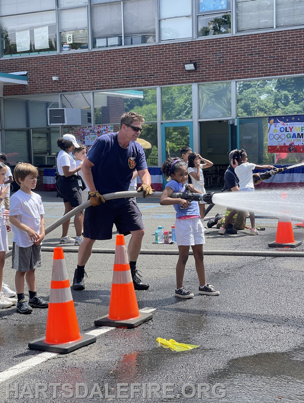 A firefighter and a child use a fire hose while others watch during a community event, with orange cones marking the area.