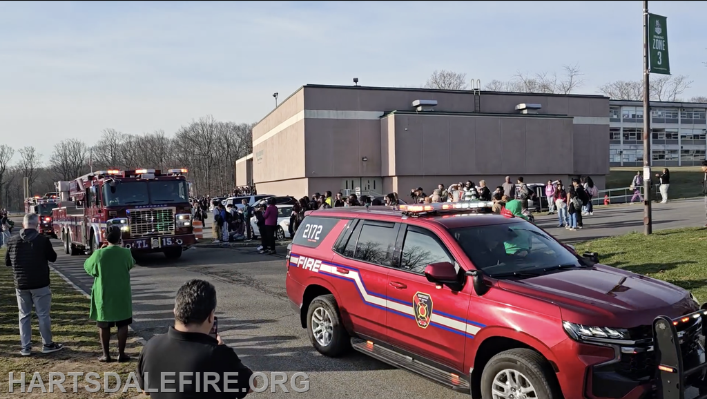 Fire trucks responding to an incident near a school, with a crowd of people gathered outside.
