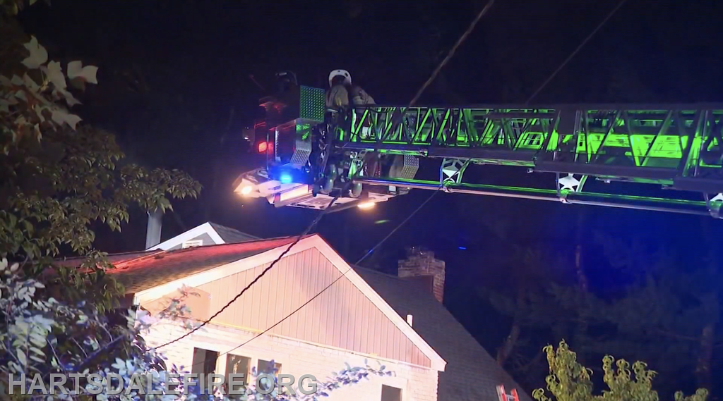 A fire truck ladder is extended towards a house at night, with firefighters and emergency lights visible.
