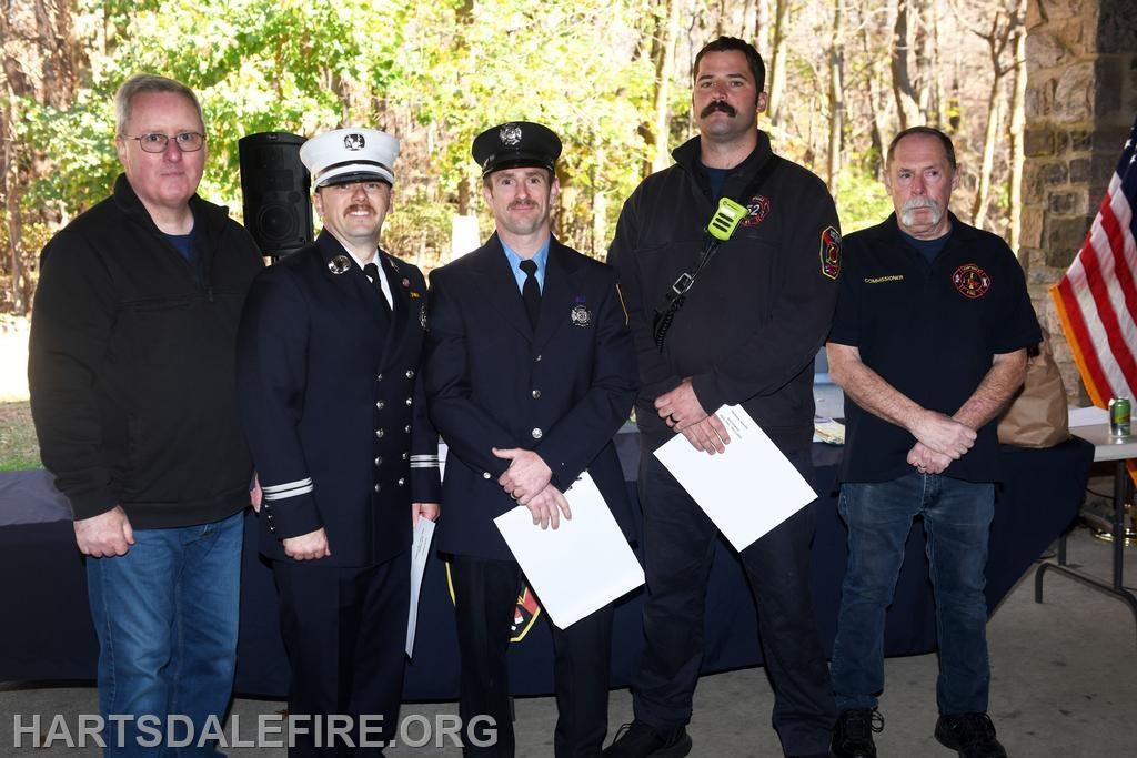 A group of five individuals, including firefighters and a commissioner, are gathered, likely for an event or ceremony outdoors.